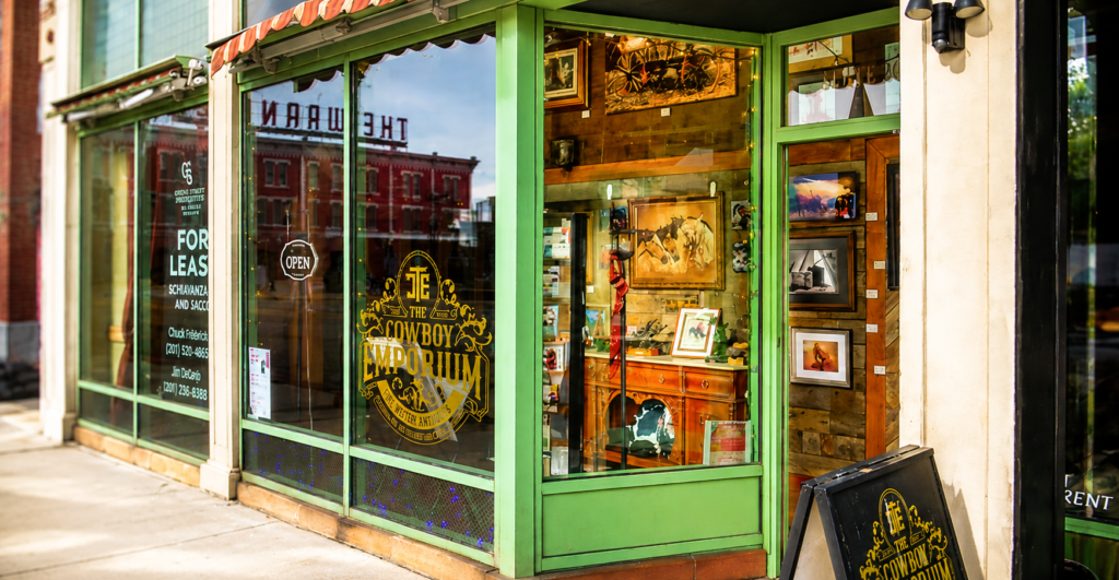 Storefront of The Cowboy Emporium in downtown Cheyenne, Wyoming, featuring large display windows, Western artwork, antiques, rustic furnishings, and a warmly lit interior visible from the sidewalk.